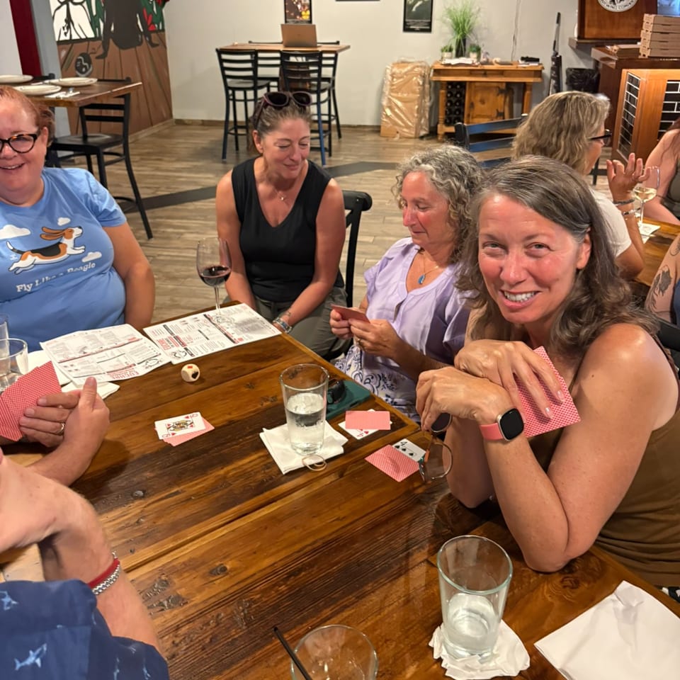Friends gather around a wooden restaurant table with menus, drinks, and playing cards, smiling during a social meal.