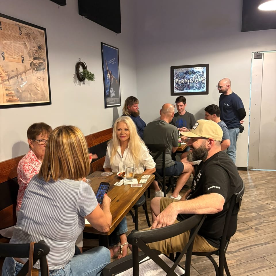 Guests socialize around tables in a warmly lit restaurant, with playing cards, drinks, wall art, and a framed Pernicious poster.