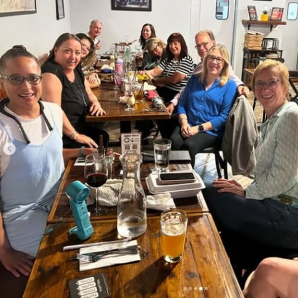 Group of friends smiling together around restaurant tables with drinks, dishes, and warm dining-room decor.