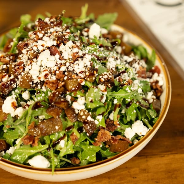 Arugula salad with crumbled feta, bacon bits, and balsamic glaze in a white bowl.