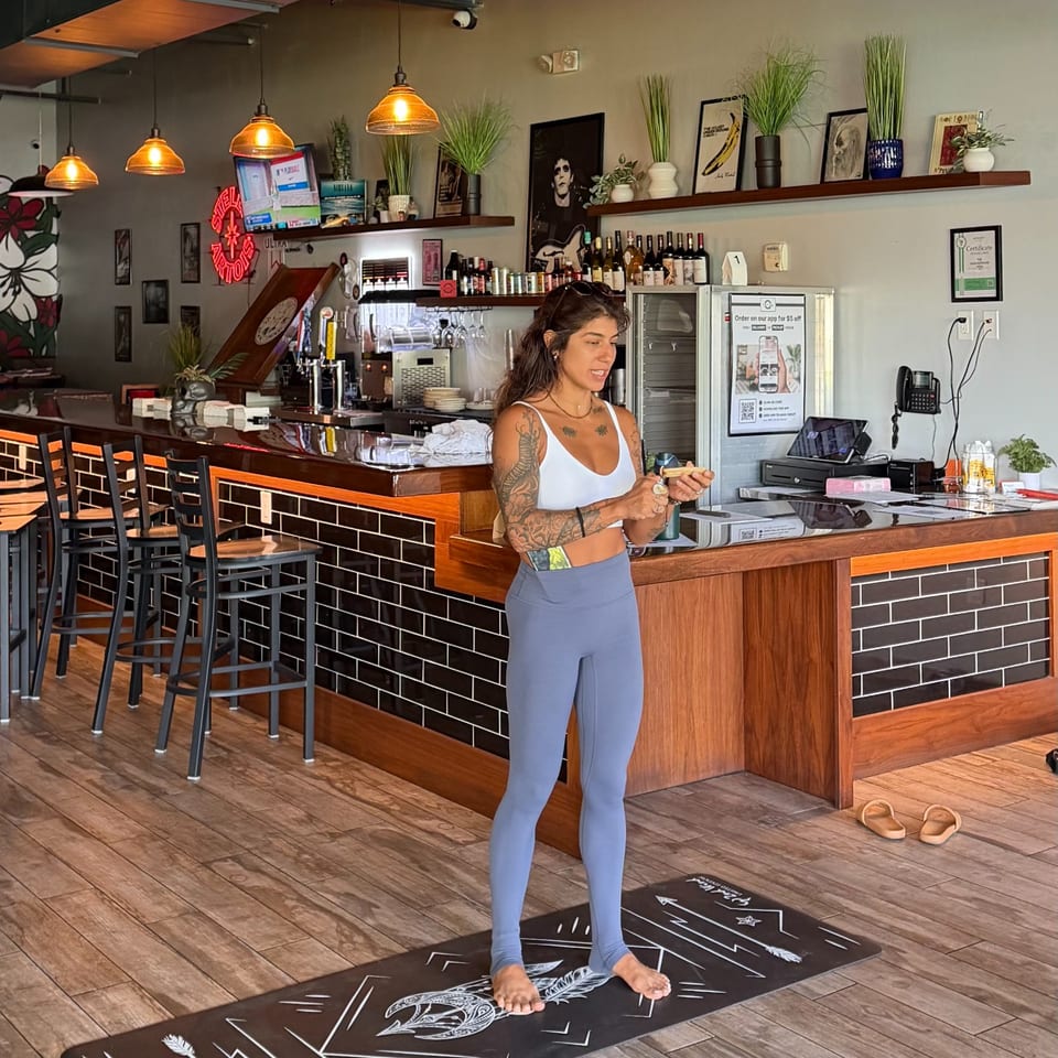 Tattooed woman stands barefoot on a yoga mat inside a warmly lit restaurant bar with pendant lights, stools, plants, and framed artwork.