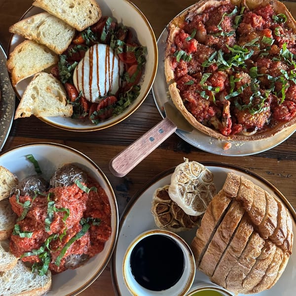 Overhead view of a deep-dish pizza, meatballs, burrata salad, and sliced bread on a wooden table.