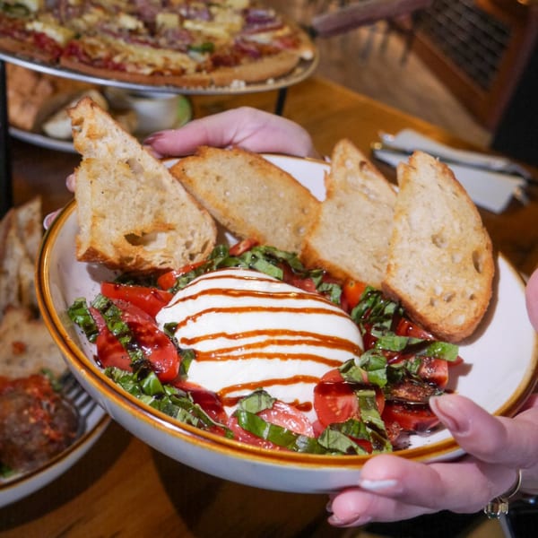 Hands holding a bowl of Caprese salad with balsamic glaze and toasted bread, pizza in background.