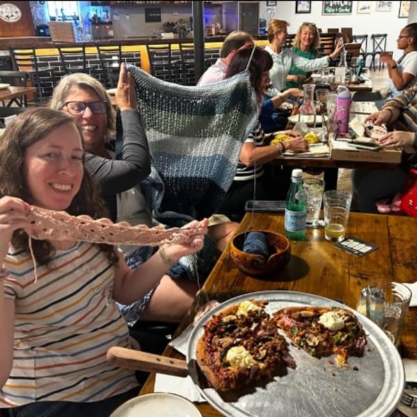 Friends dine together at a lively restaurant table, with drinks, food, and a large plate of sandwiches in the foreground.