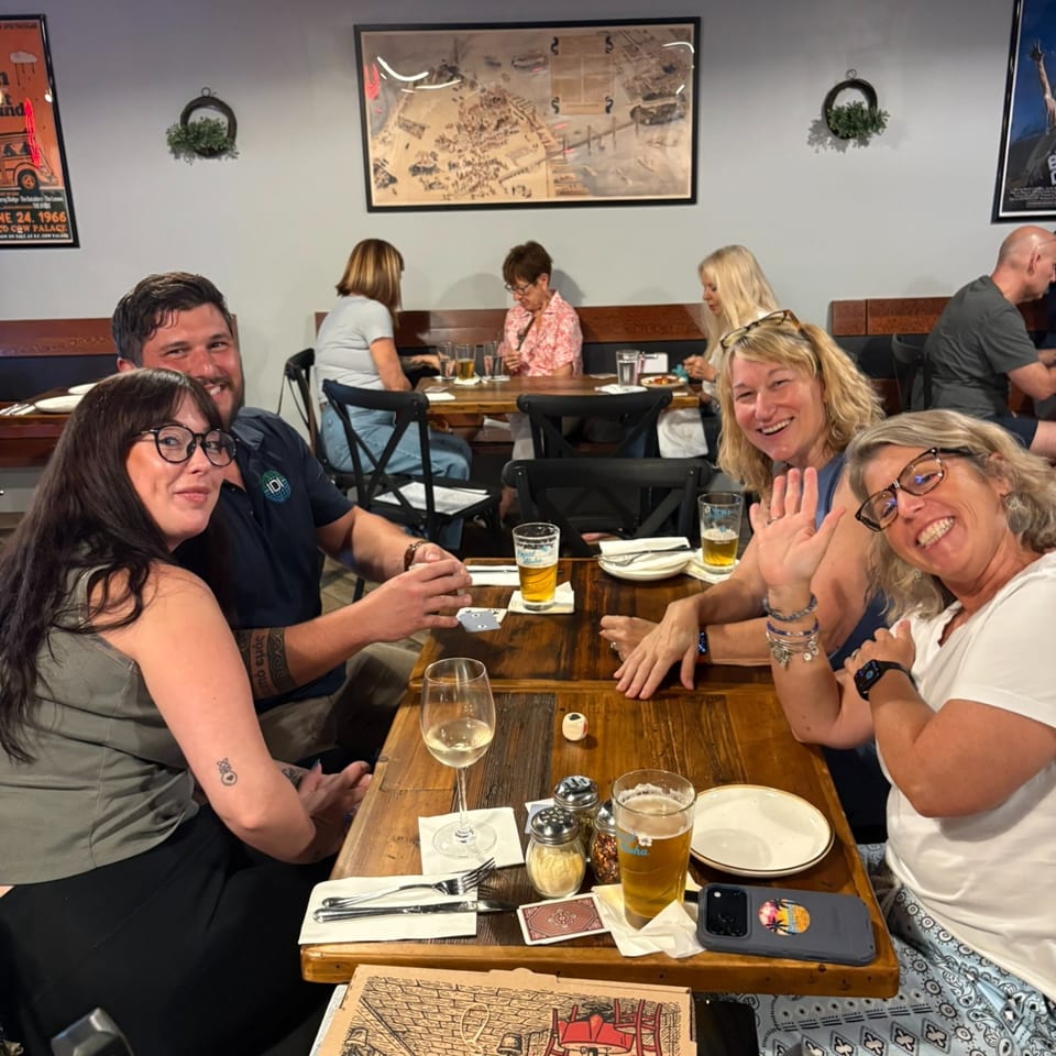 Friends smile and wave around a wooden restaurant table set with plates, silverware, wine, and beer.